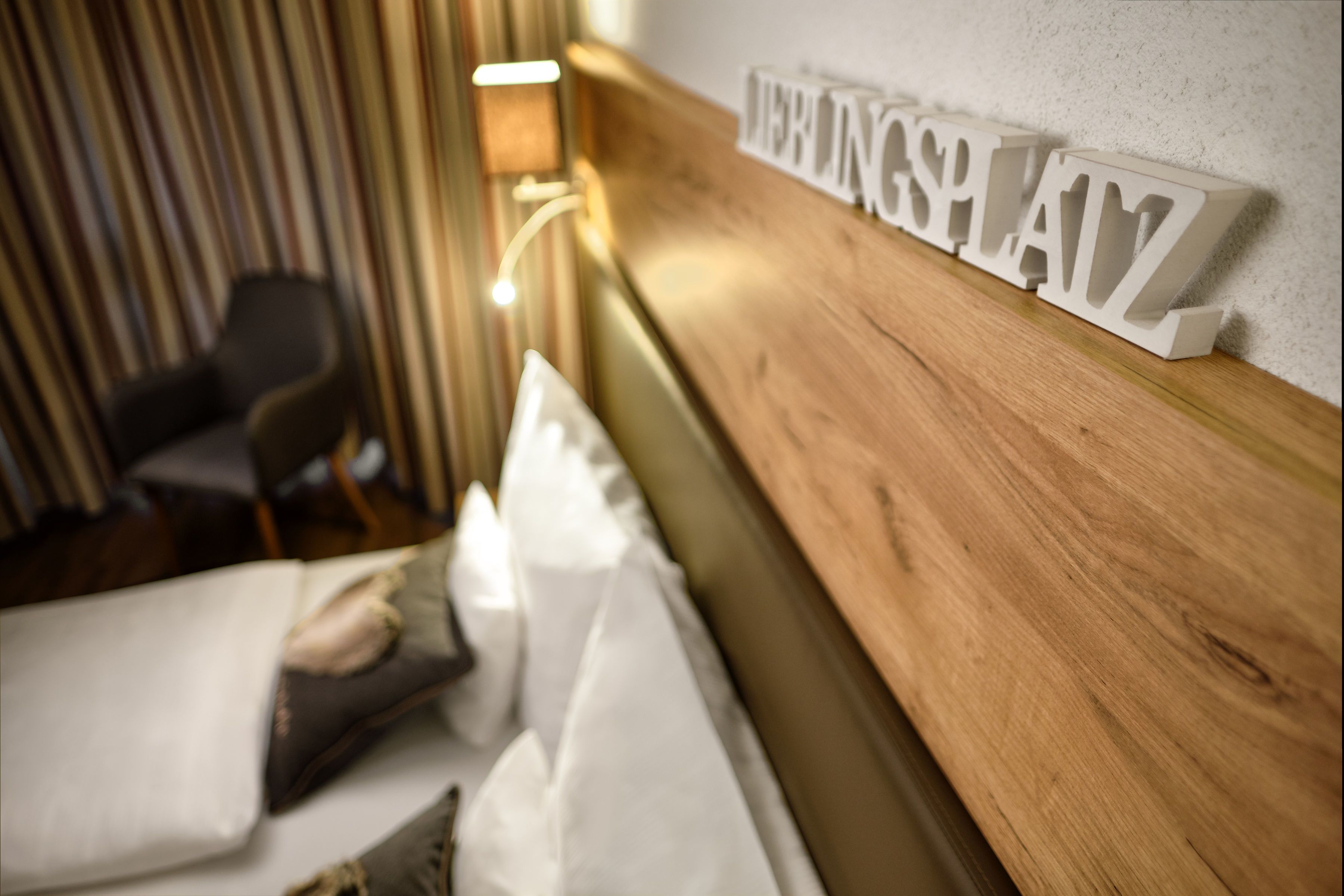Een hotelkamer met een houten hoofdeinde, een bed met witte lakens, en een bordje aan de muur waarop 'HEINZ PLAYZ' staat.