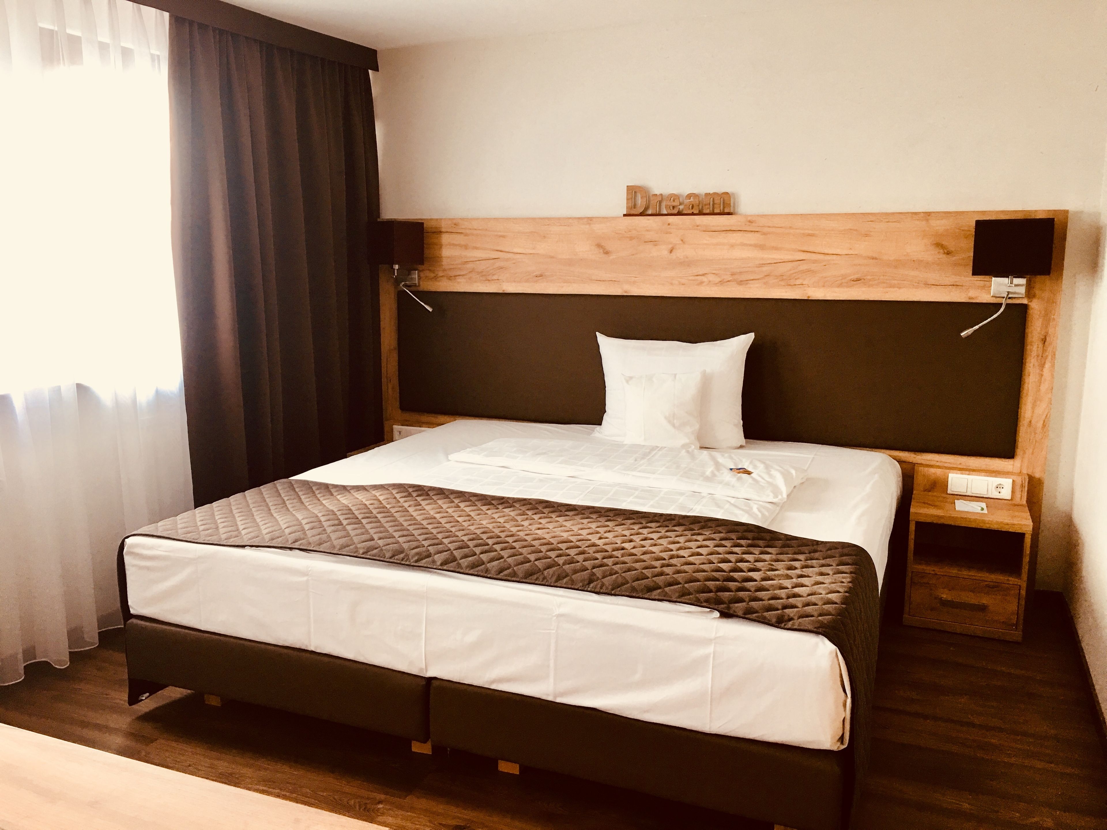 A neatly made bed with a quilted cover, a wooden headboard, a bedside table with a power outlet, and a wall-mounted speaker.