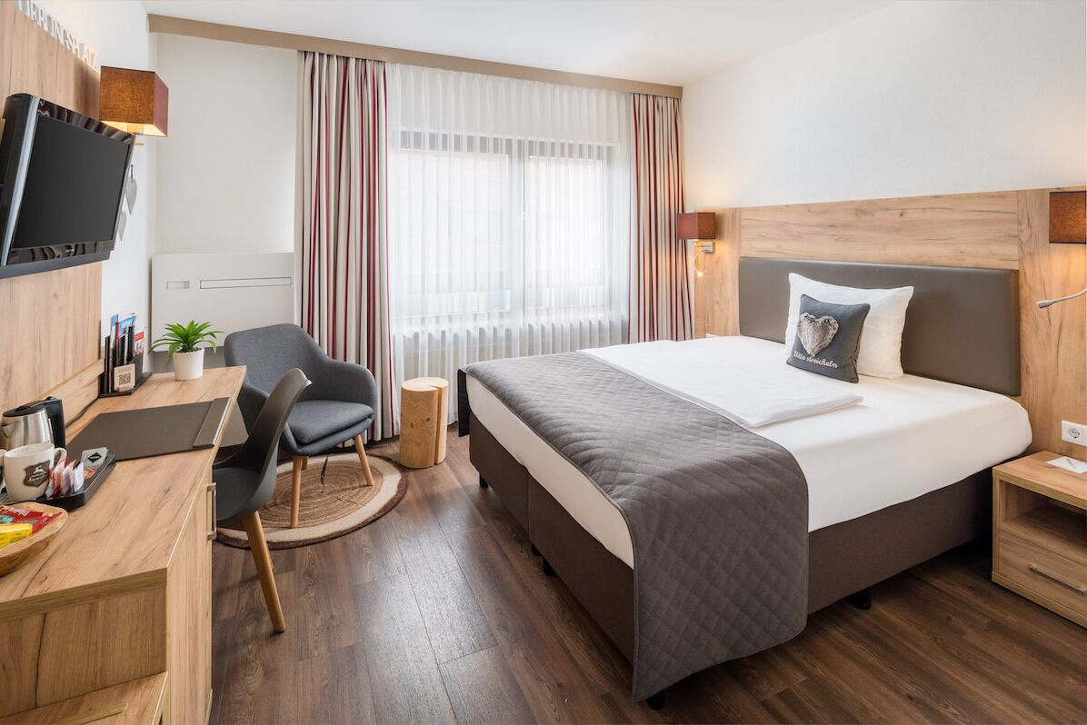 A modern hotel room with a bed, a desk, a chair, a TV, and a window with curtains.