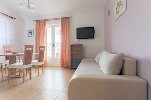 A living room with a sofa, a TV mounted on the wall, a dining table with chairs, and a view through a glass door. - Apartmani Goic (Jelsa)