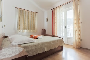 A bedroom with a bed, a wooden nightstand, a chair, and a window with curtains. - Apartmani Goic (Jelsa)