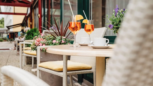 A round table with wicker chairs, a white cup, and two glasses of a beverage with a straw and an orange slice. - Kurhotel Panland (Bad Fuessing)