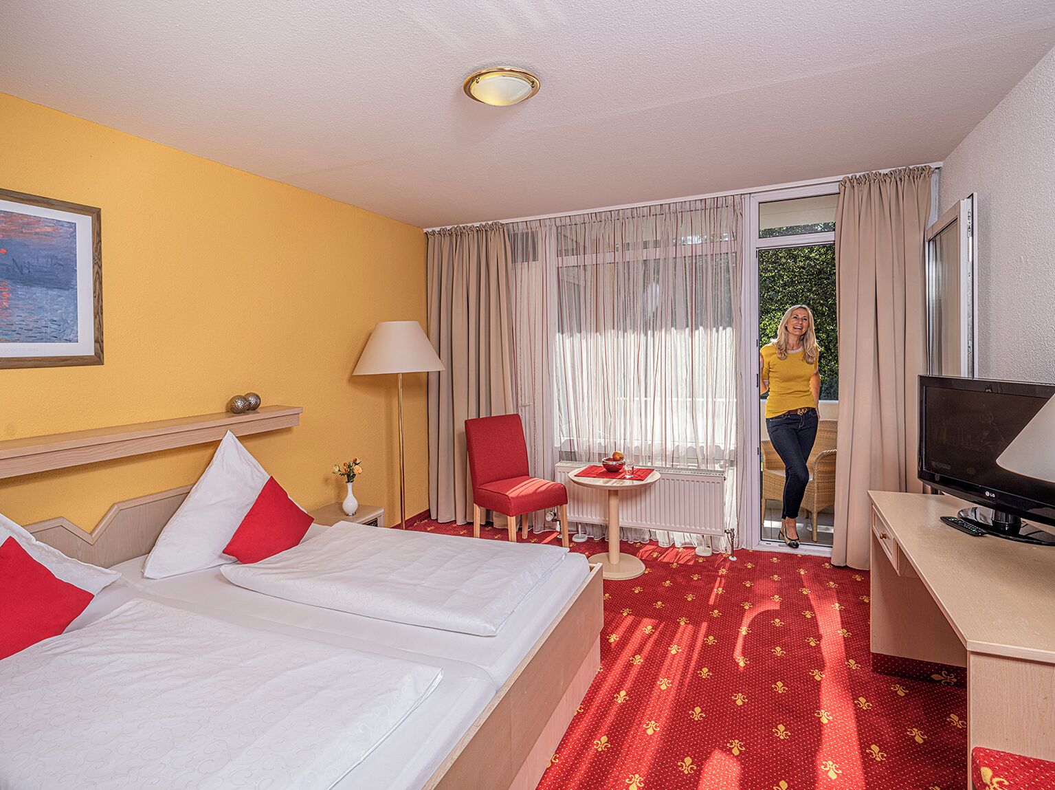 A hotel room with two beds, a desk with a TV, a small table with a red bowl, a chair, and a window with curtains.