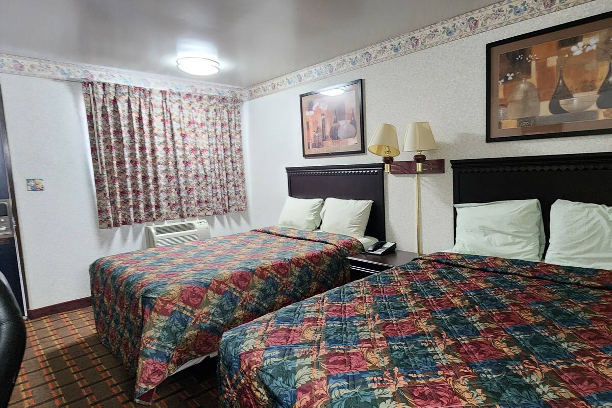 Une chambre d’hôtel avec deux lits, un bureau, une chaise, une fenêtre avec des rideaux à motifs floraux, un tableau encadré au mur et une table de chevet avec des lampes.