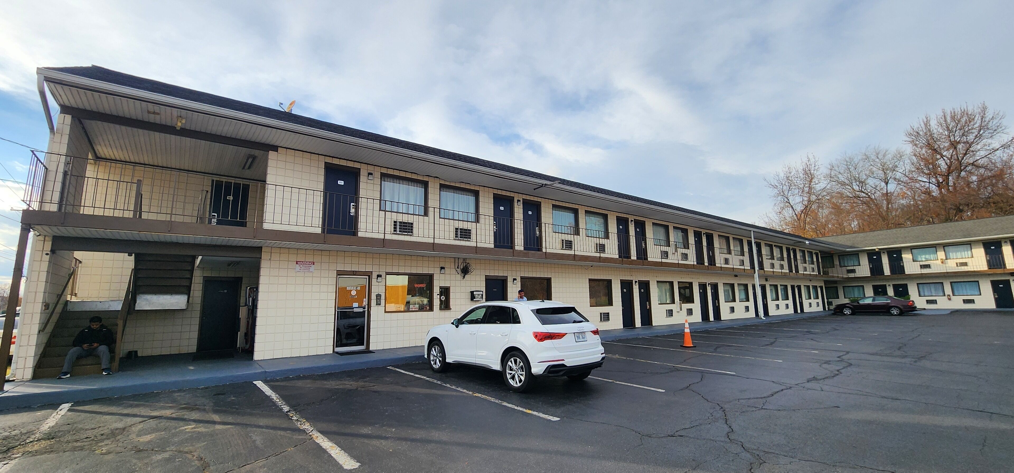 A multi-story motel with a parking lot, a white SUV parked in front, and a person sitting on the steps.