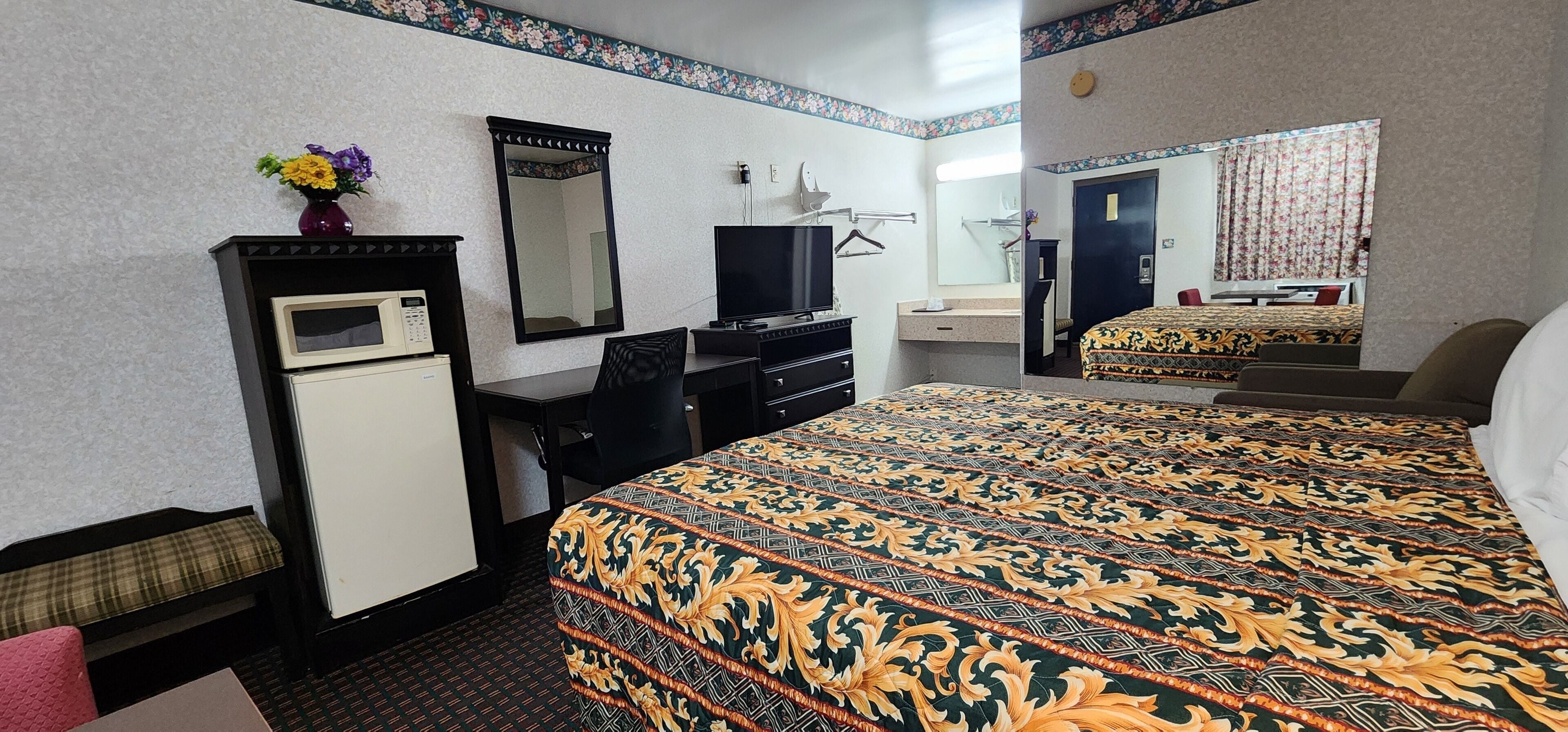 A hotel room with a bed, a desk with a computer, a TV, a mirror, and a small fridge.