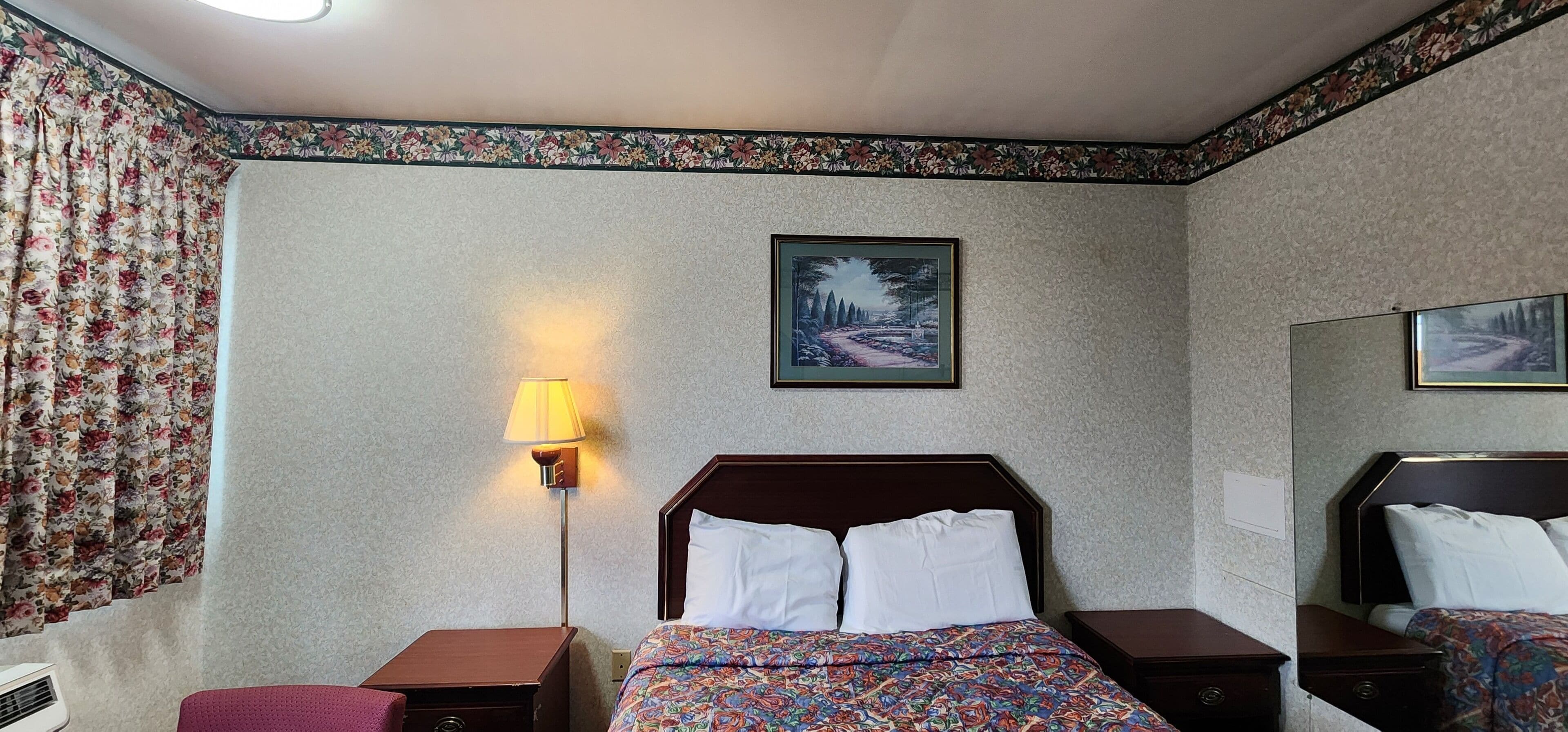 A hotel room with a bed, a nightstand, a lamp, a floral curtain, and a framed picture on the wall.