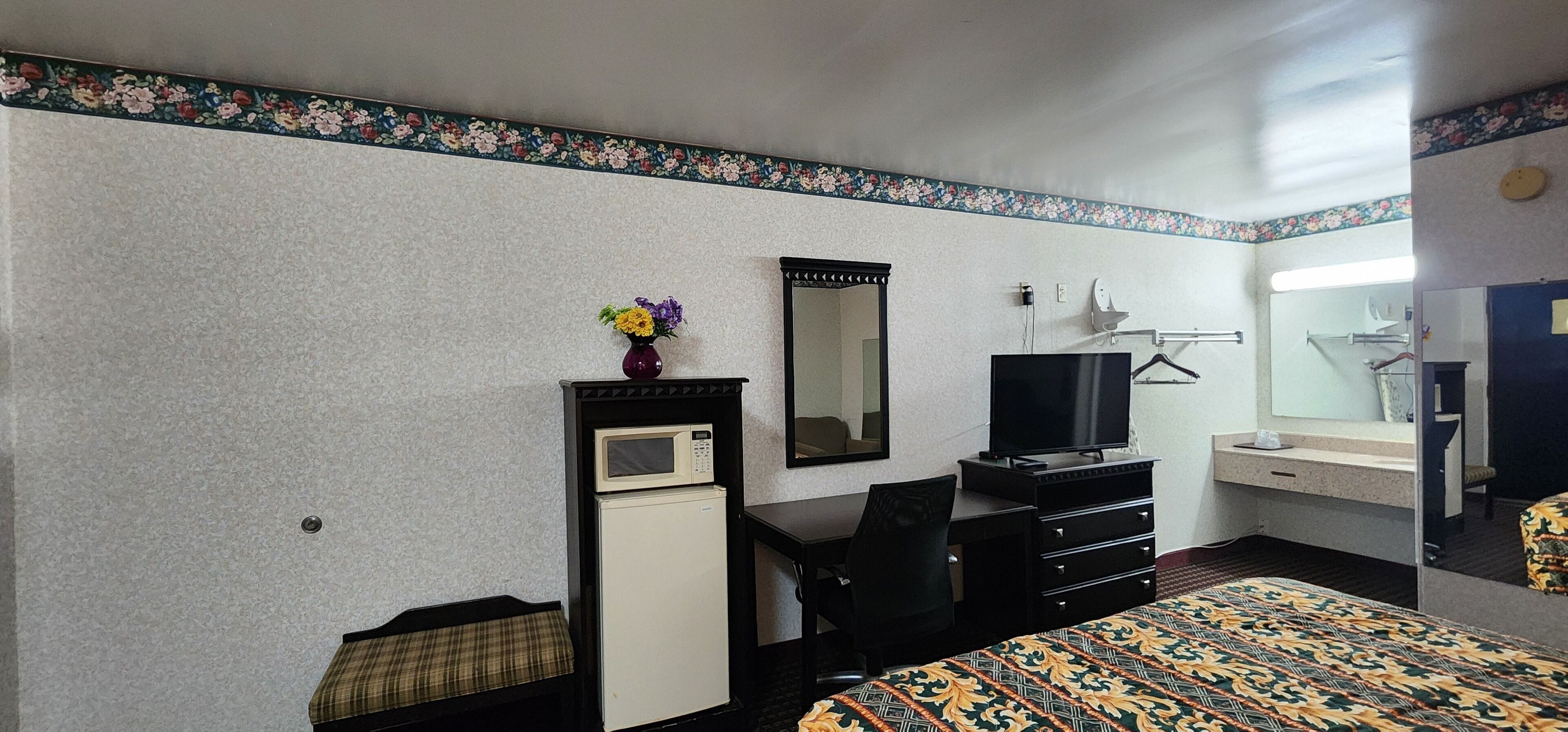 A hotel room with a bed, a desk with a television, a small refrigerator, a mirror, and a wall-mounted TV.