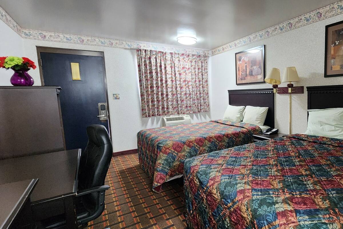 Une chambre d’hôtel avec deux lits, un bureau, une chaise, un réfrigérateur, une fenêtre avec des rideaux à motifs floraux et une lampe fixée au mur.