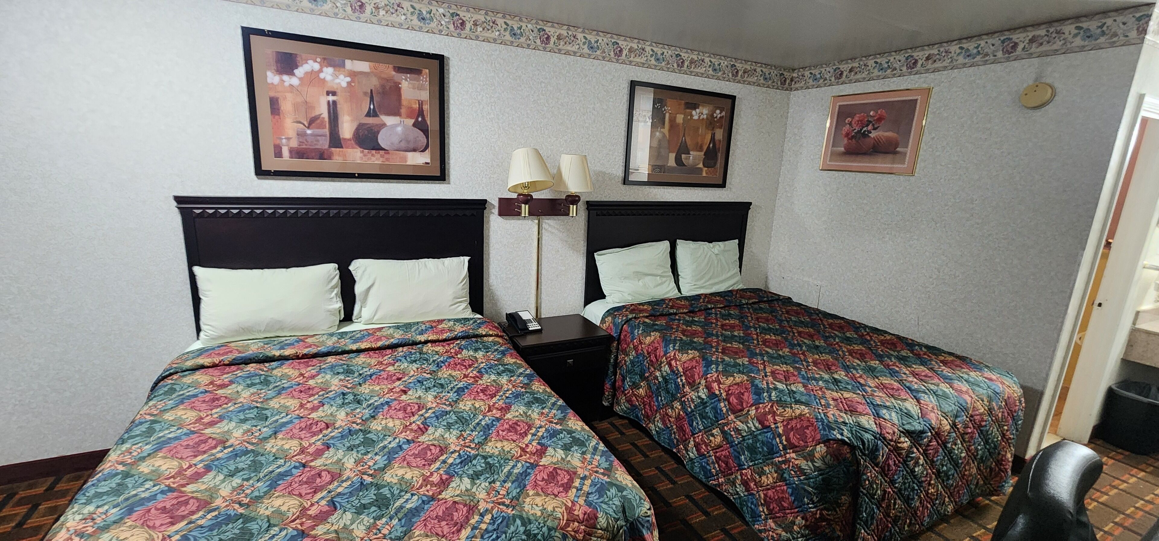 A hotel room with two beds, each with a floral bedspread and a dark headboard. There are two wall-mounted lamps and framed artwork above the beds.