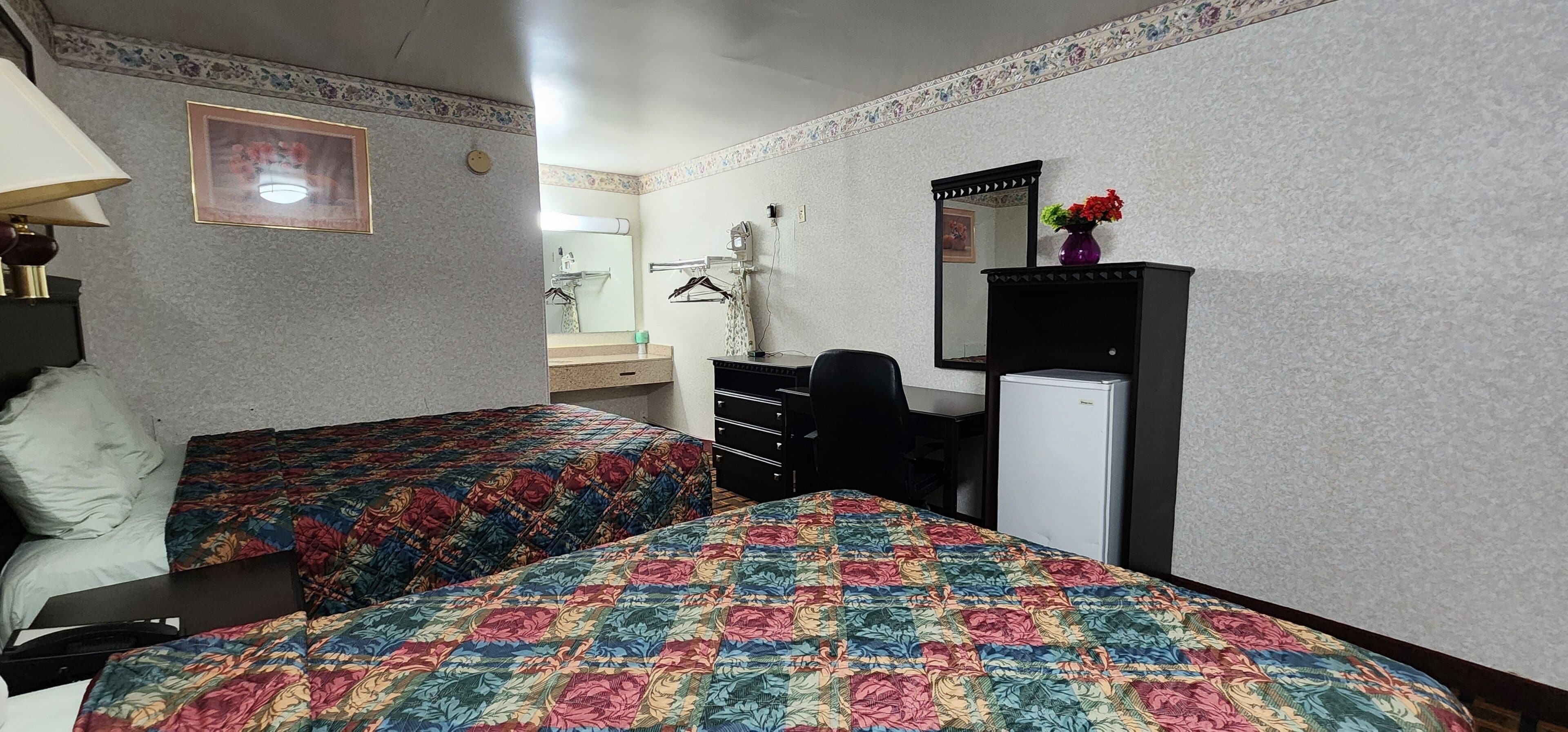 A hotel room with two beds, a desk, a mirror, a dresser, and a refrigerator.
