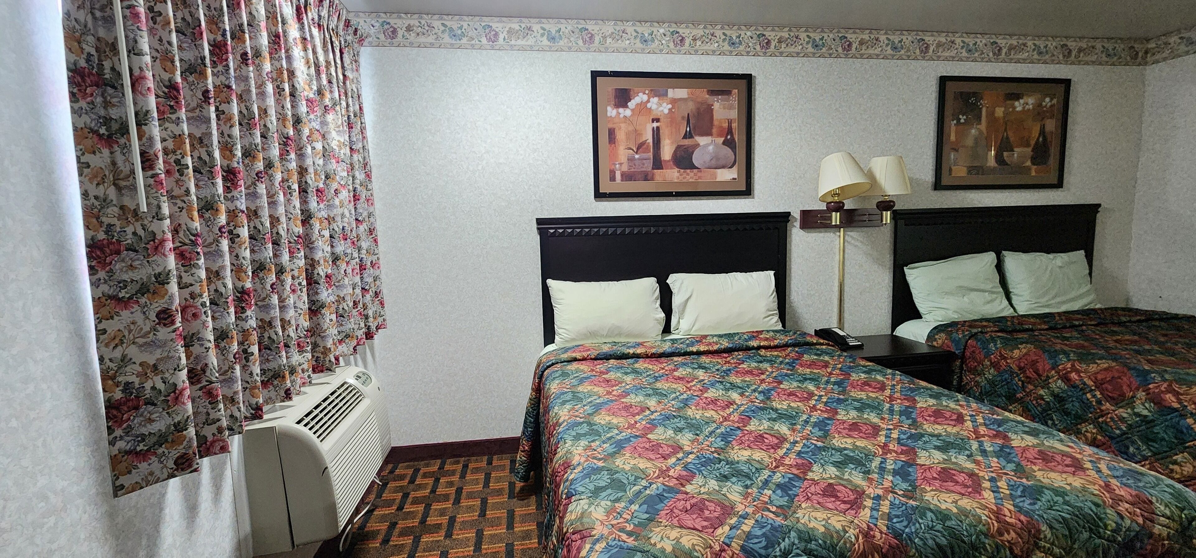 A hotel room with two beds, floral curtains, a radiator, and two wall-mounted lamps.