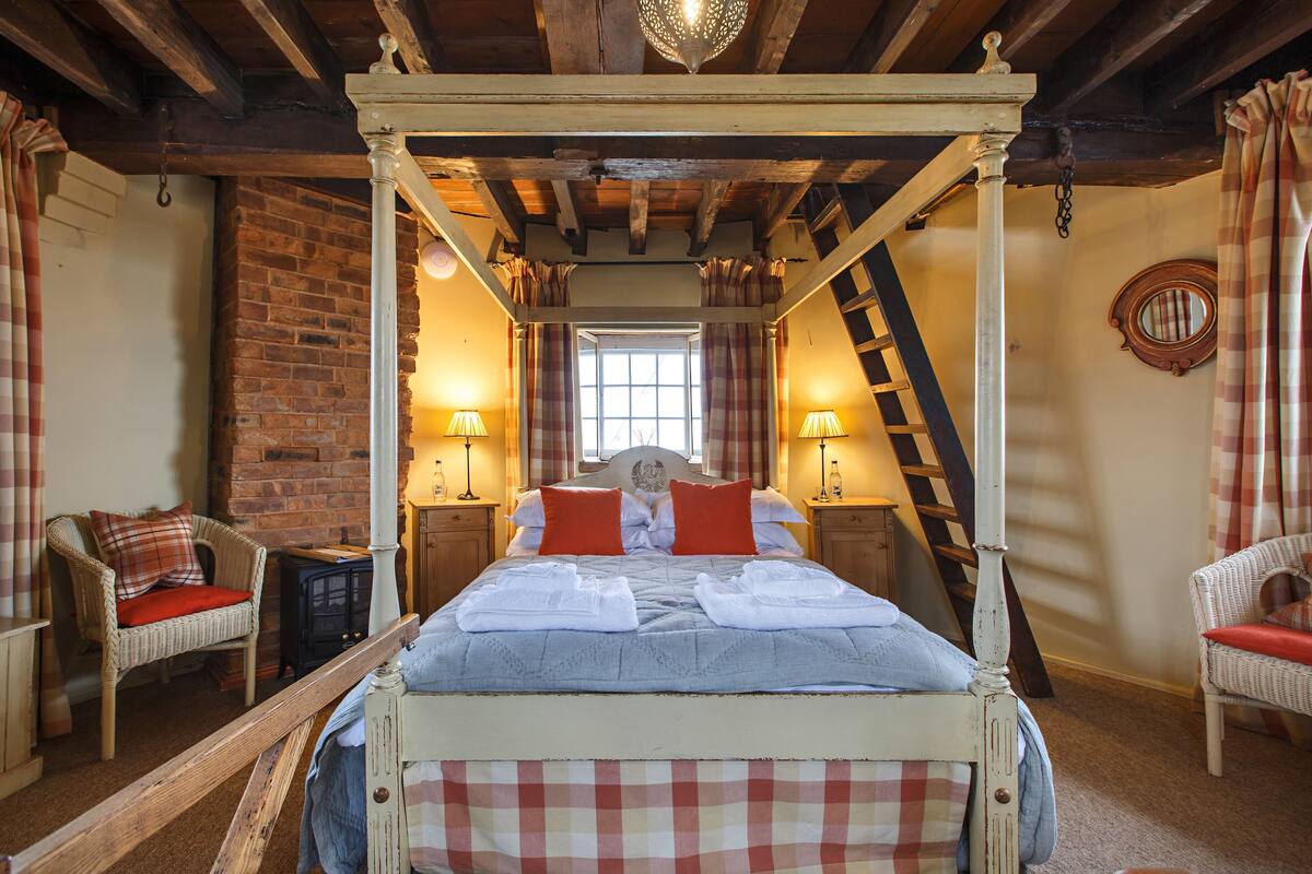 A four-poster bed with a checkered bedspread, two bedside lamps, a brick wall, a window, and a mirror on the wall.