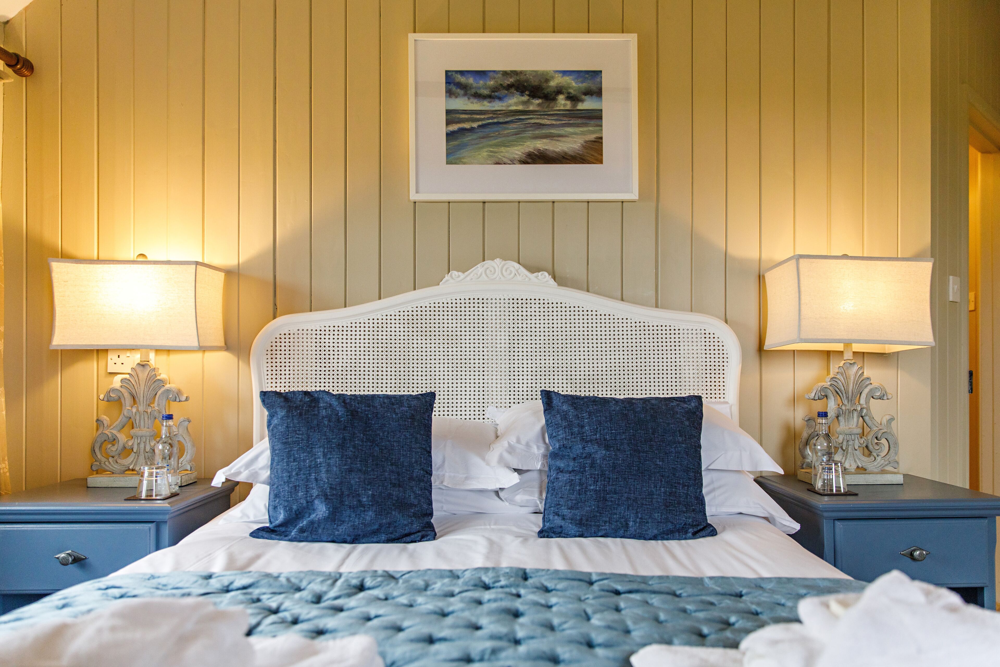 A neatly made bed with a white headboard, two blue pillows, and a blue bedspread. There are two bedside tables with lamps and decorative items.