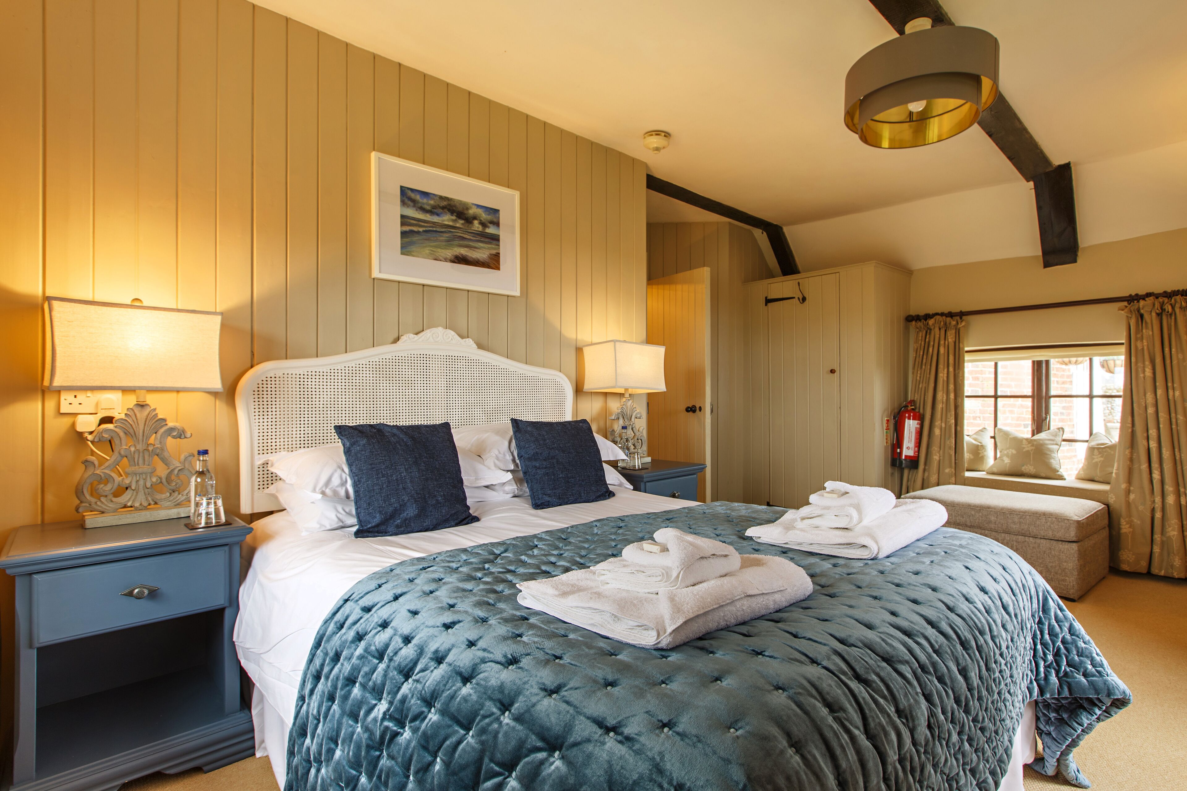 A well-appointed hotel room with a large bed, two bedside tables, a nightstand, a window with curtains, and a painting on the wall.