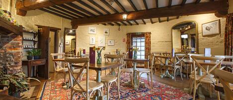 A rustic dining area with wooden tables and chairs, a patterned rug, and a brick wall.