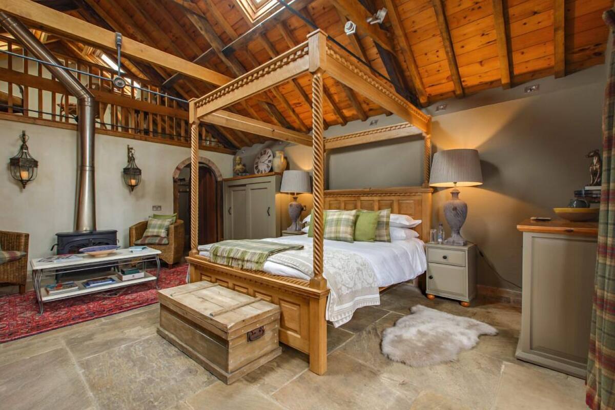 A rustic bedroom with a four-poster bed, wooden furniture, a stone fireplace, and a stone floor.