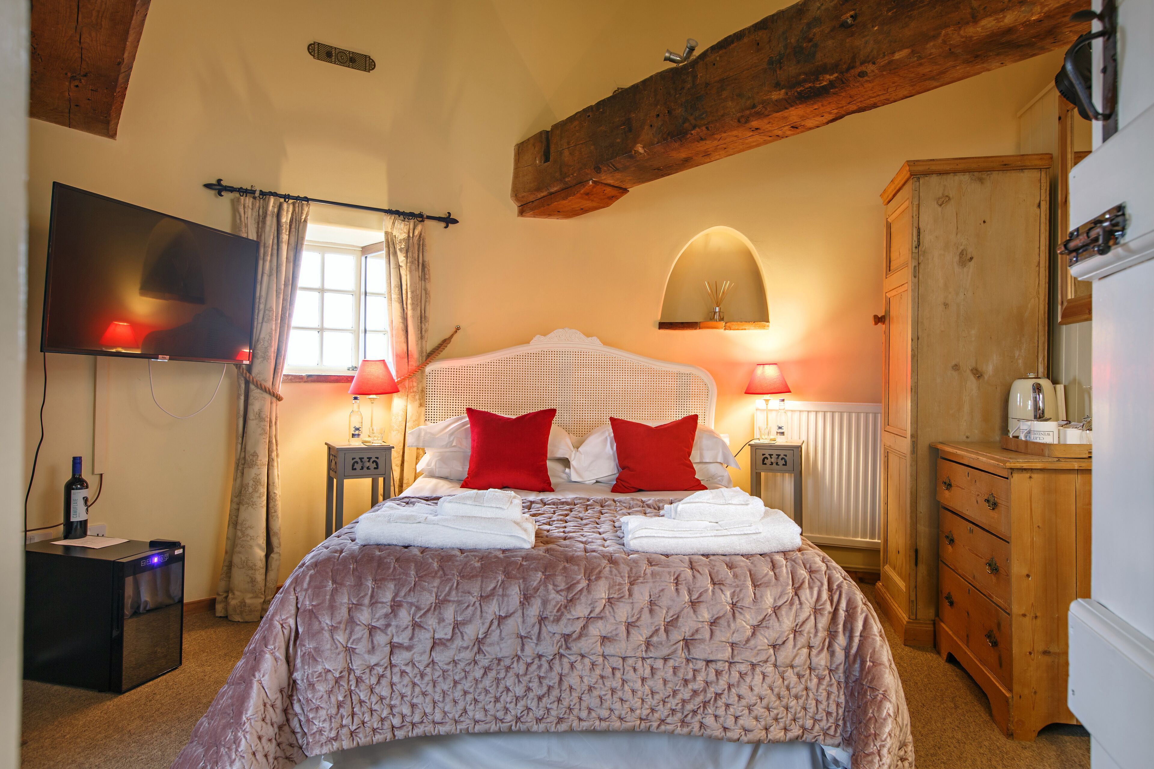 Ein ordentlich bezogenes Bett mit einem gemusterten Überwurf und roten Kissen, ein Flachbildfernseher an der Wand, eine hölzerne Kommode und ein Holzbalken an der Decke.