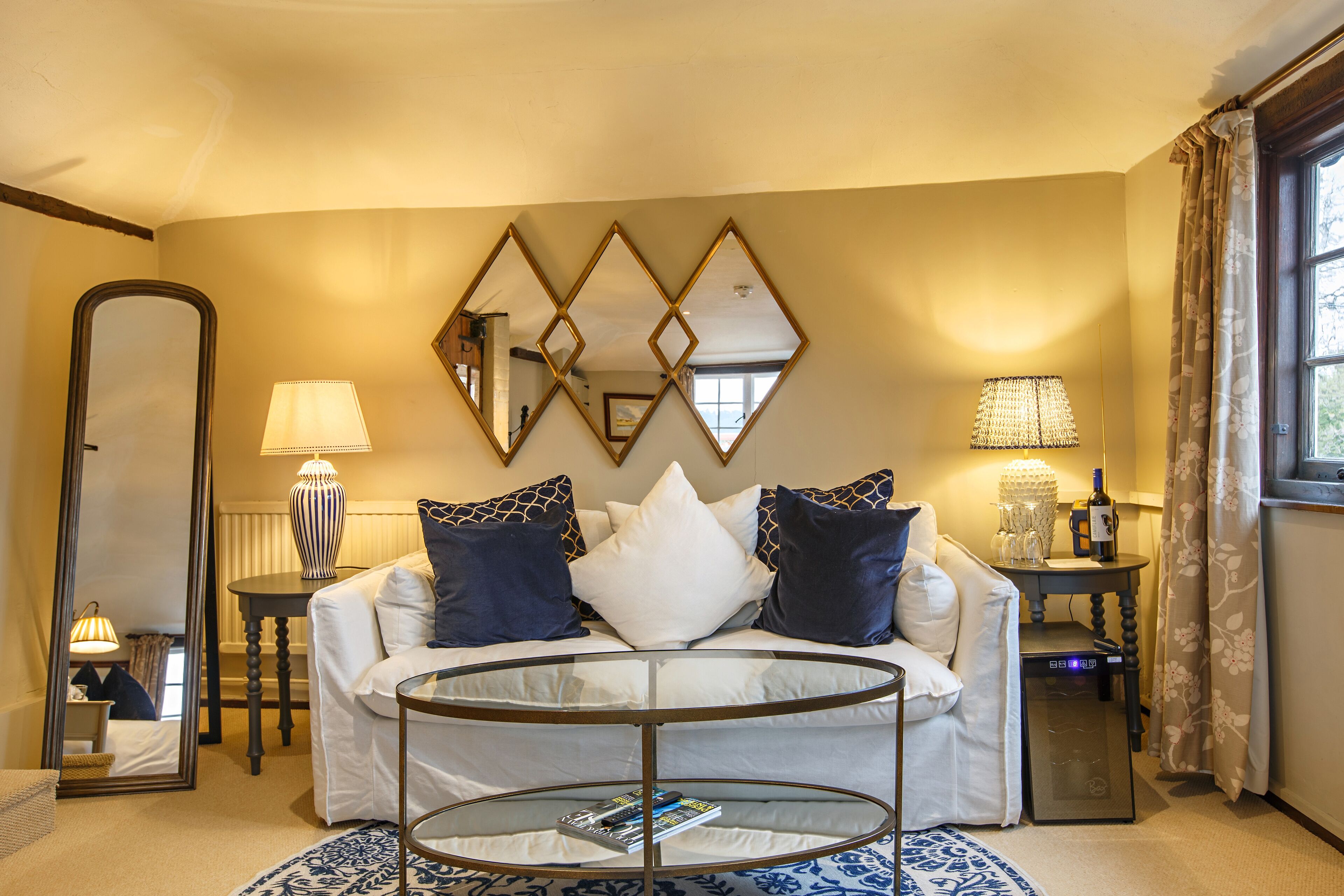 Un salon avec une table basse ronde en verre, un canapé agrémenté de coussins à motifs, un grand miroir et une lampe posée sur une table d’appoint.