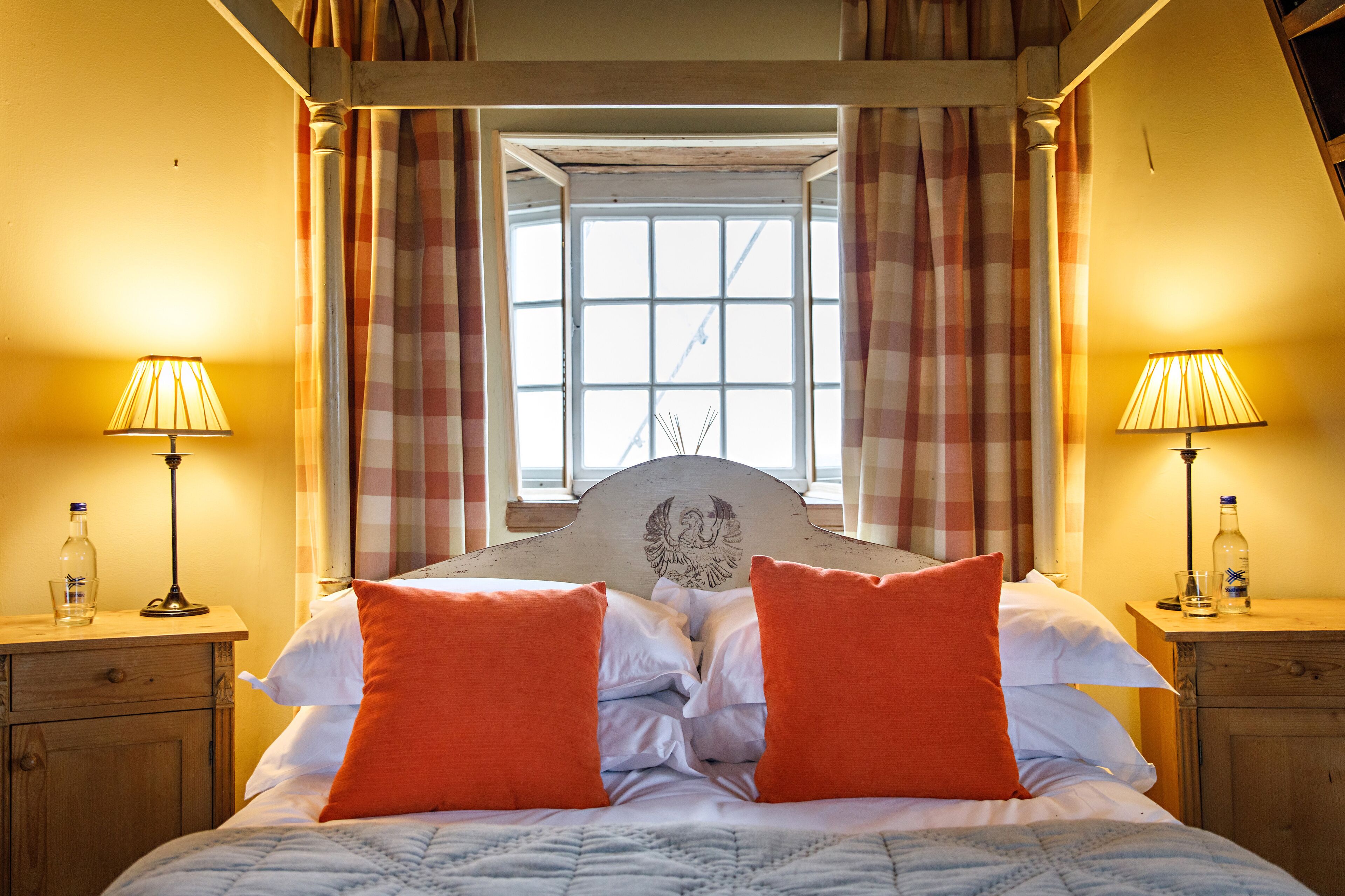 A bed with two orange pillows, a wooden headboard with an eagle design, and a window with checkered curtains.