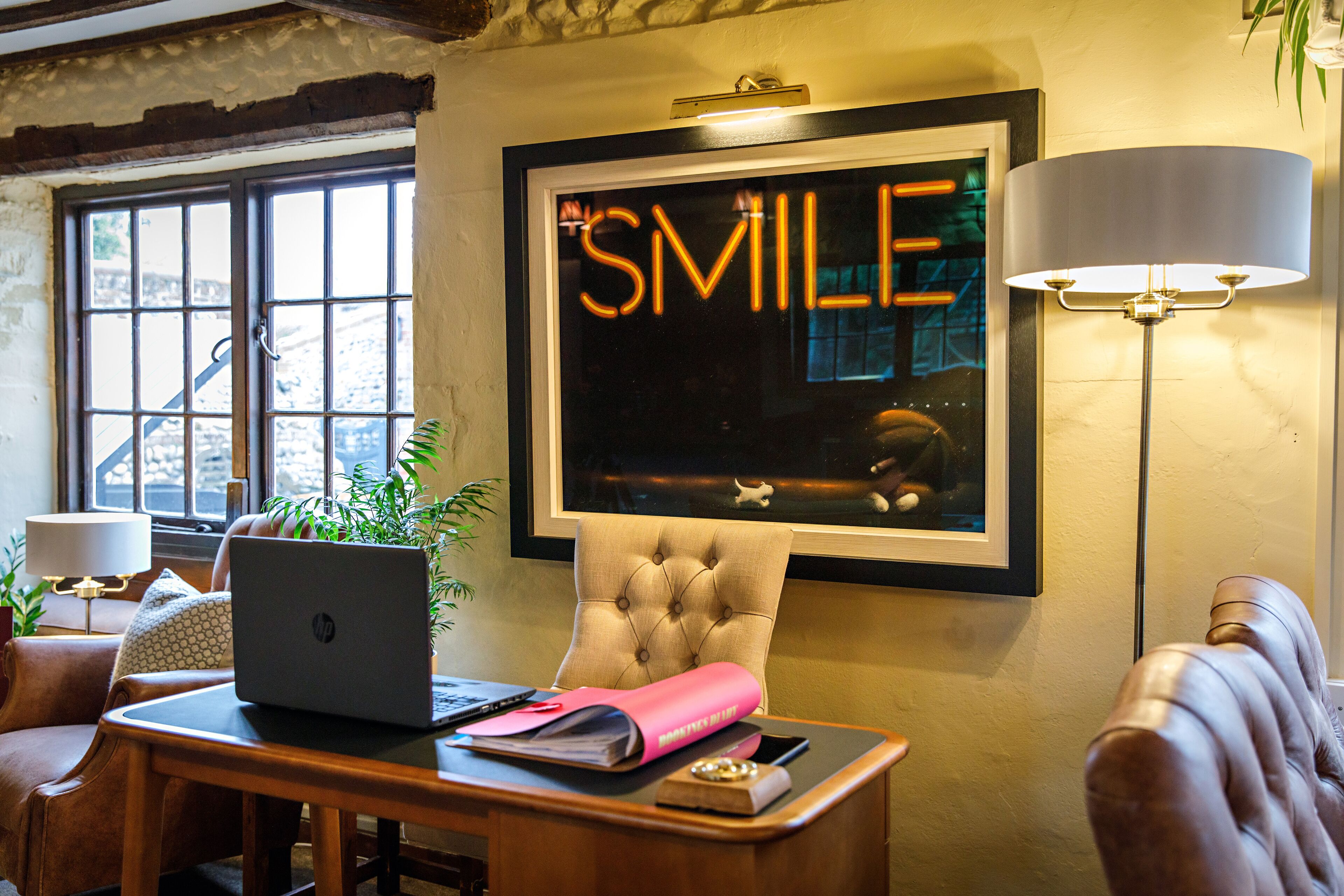Um quarto com um laptop sobre a mesa, um quadro com a palavra 'SMILE' na parede, uma luminária e uma cadeira.