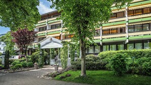A hotel with a green and white facade, a sign reading "Unter Den Linden," and a well-maintained garden with a pathway leading to the entrance. - Kurhotel Unter den Linden (Bad Fuessing)
