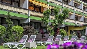 A hotel with multiple balconies, outdoor seating with cushions, and various trees and shrubs. - Kurhotel Unter den Linden (Bad Fuessing)