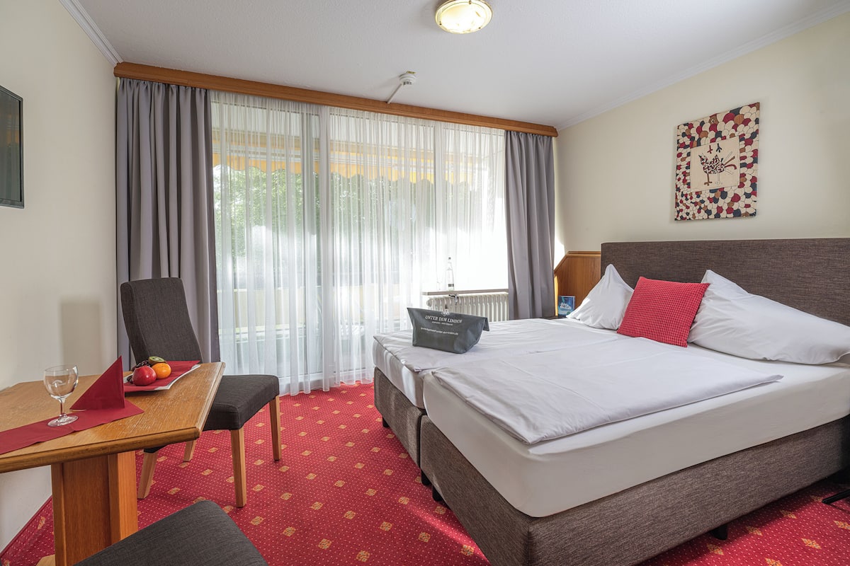 A hotel room with a bed, a desk with a chair, a table with a glass of wine, and a window with curtains.