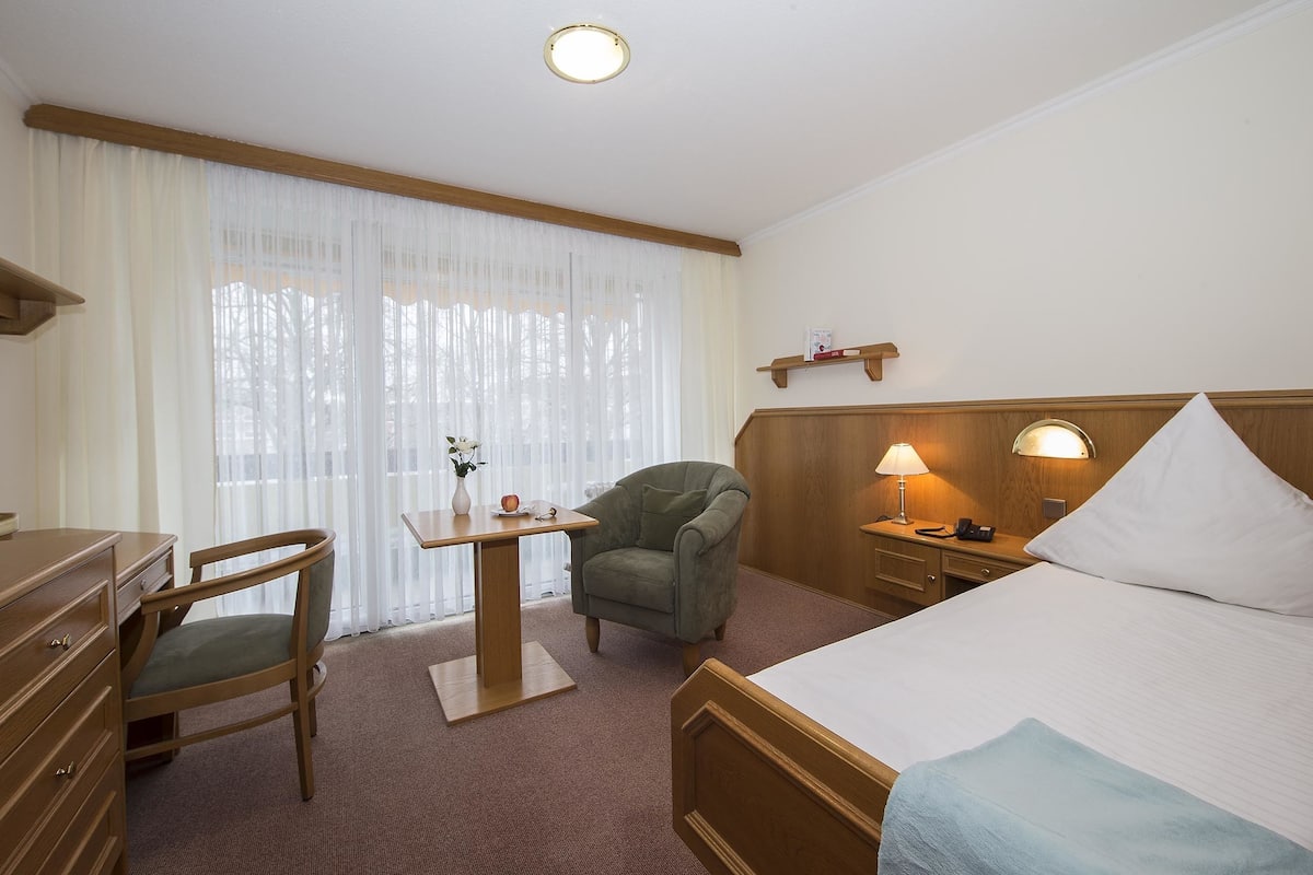 A hotel room with a bed, a desk, a chair, a nightstand, and a lamp.