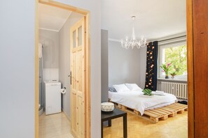 A bedroom with a wooden bed, a small bedside table, a chandelier, and a window with curtains. - Apartament Targi MTP (Poznan)