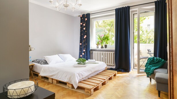 Een slaapkamer met een bed op houten pallets, een kroonluchter, een raam met gordijnen, een radiator en uitzicht op het groen.