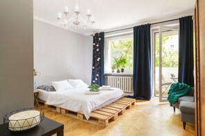 A bedroom with a bed on wooden pallets, a chandelier, a window with curtains, a radiator, and a view of the outdoors. - Apartament Targi MTP (Poznan)