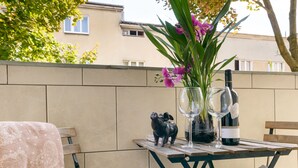 Een terras met een tafel gedekt voor twee personen, compleet met een fles wijn, twee glazen en een vaas met bloemen. Er staat een stoel met een plaid en een stoel met een kussen.