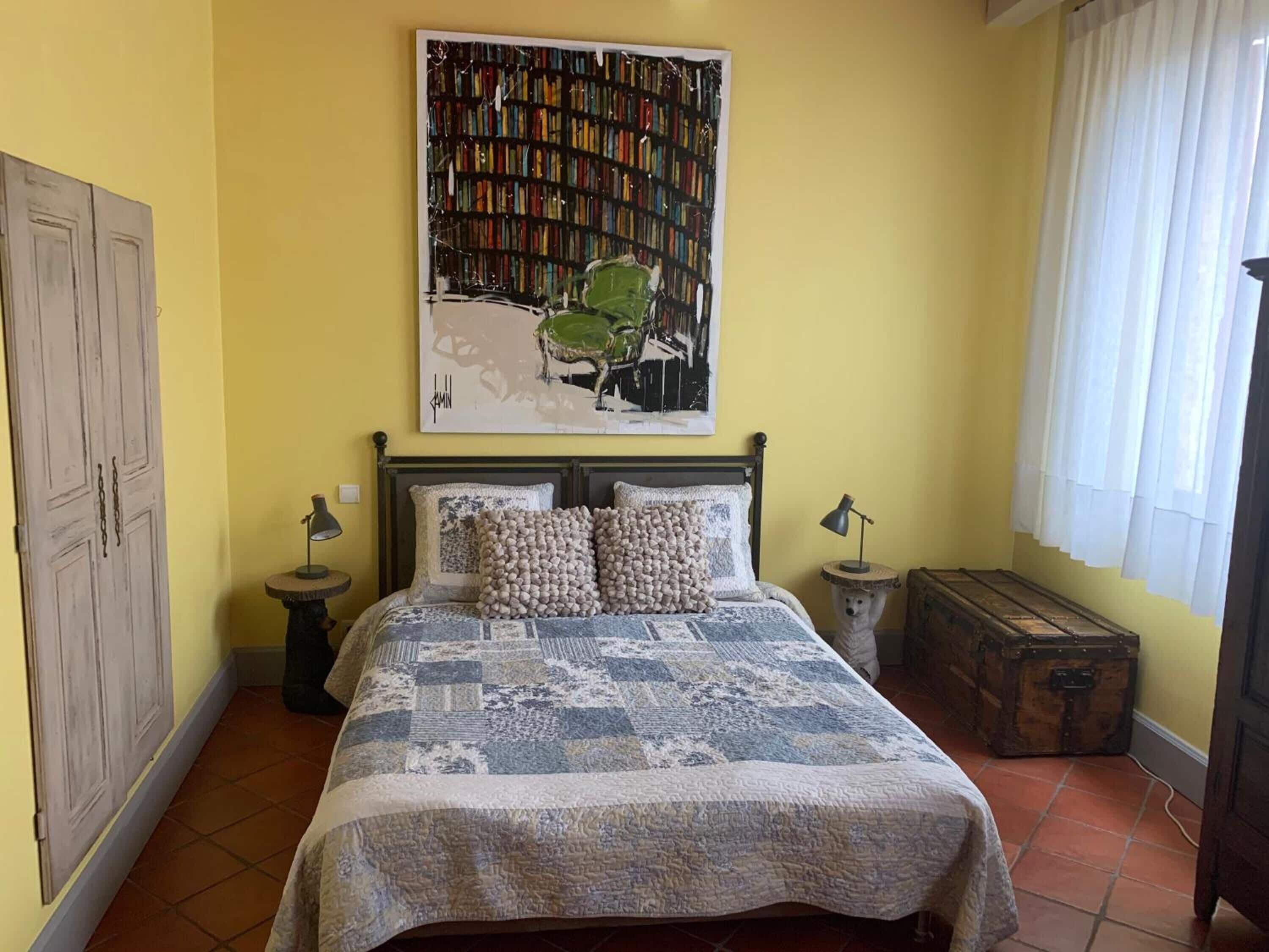 Un dormitorio con cama, estantería, ventana con cortinas, armario y una pequeña mesa redonda.