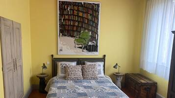 Un dormitorio con cama, estantería, ventana con cortinas, armario y una pequeña mesa redonda.