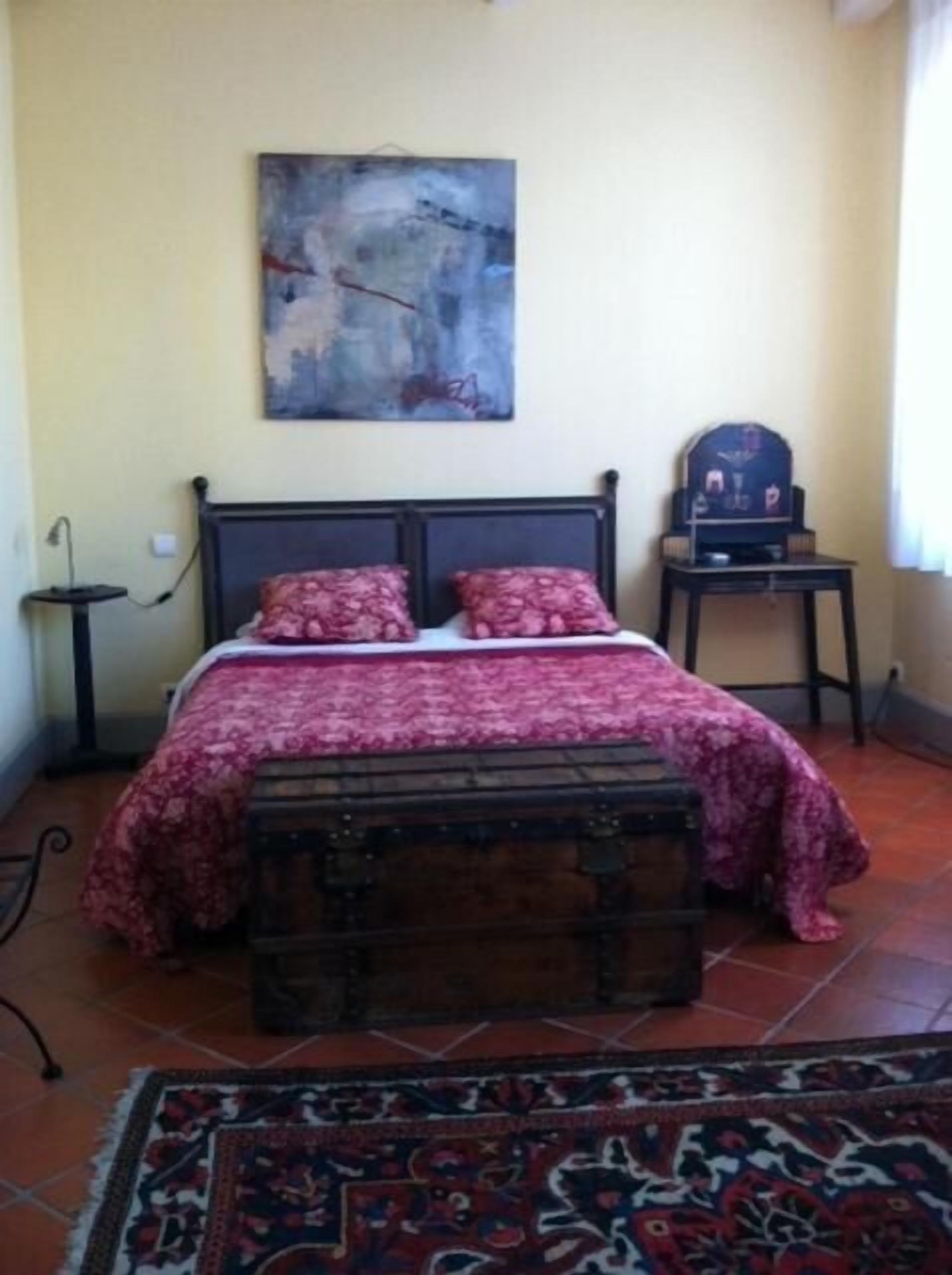 Un dormitorio con una cama, una mesita de noche, una pequeña mesa, un banco, un cuadro y una alfombra.