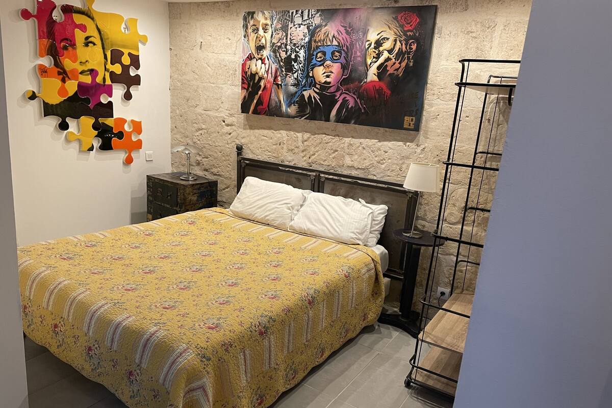 Quarto com cama, mesa de cabeceira, abajur, uma obra de arte na parede e um painel decorativo de quebra-cabeça.