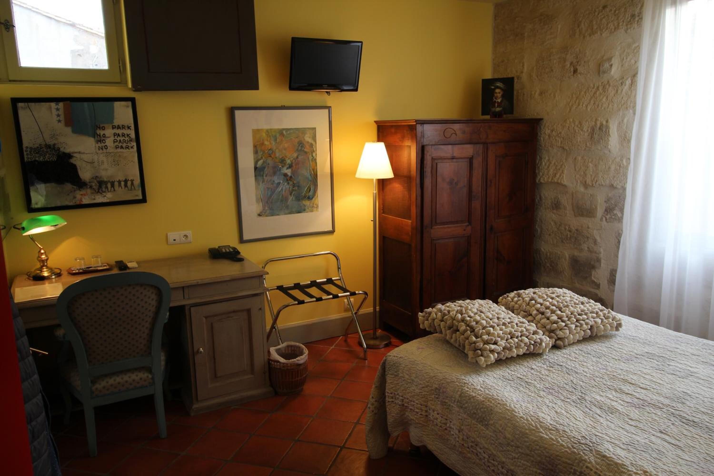 A bedroom with a bed, a desk, a chair, a lamp, a TV, and artwork on the walls.