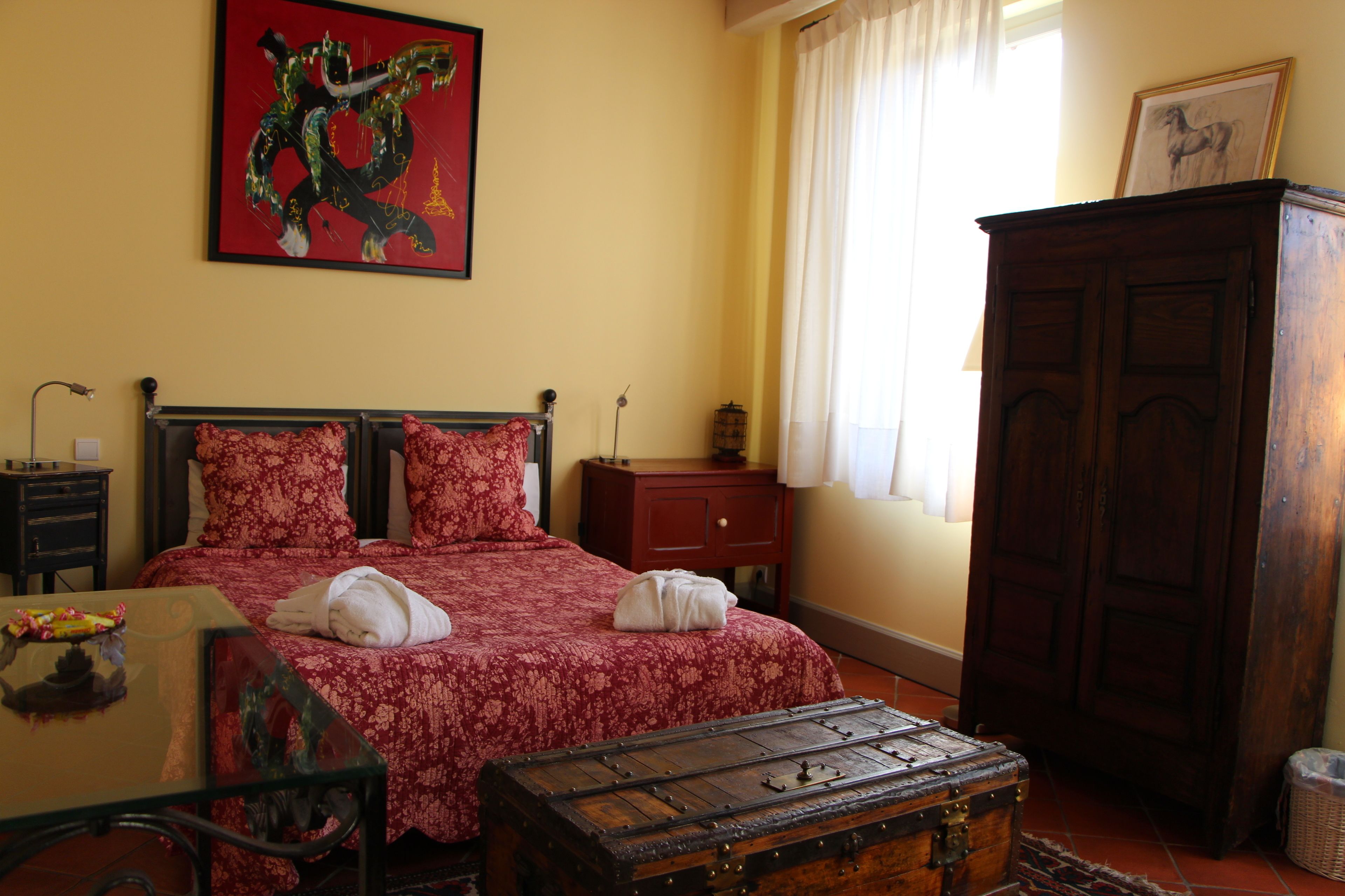 A bedroom with a bed, a wooden chest, a nightstand, a lamp, and a painting on the wall.