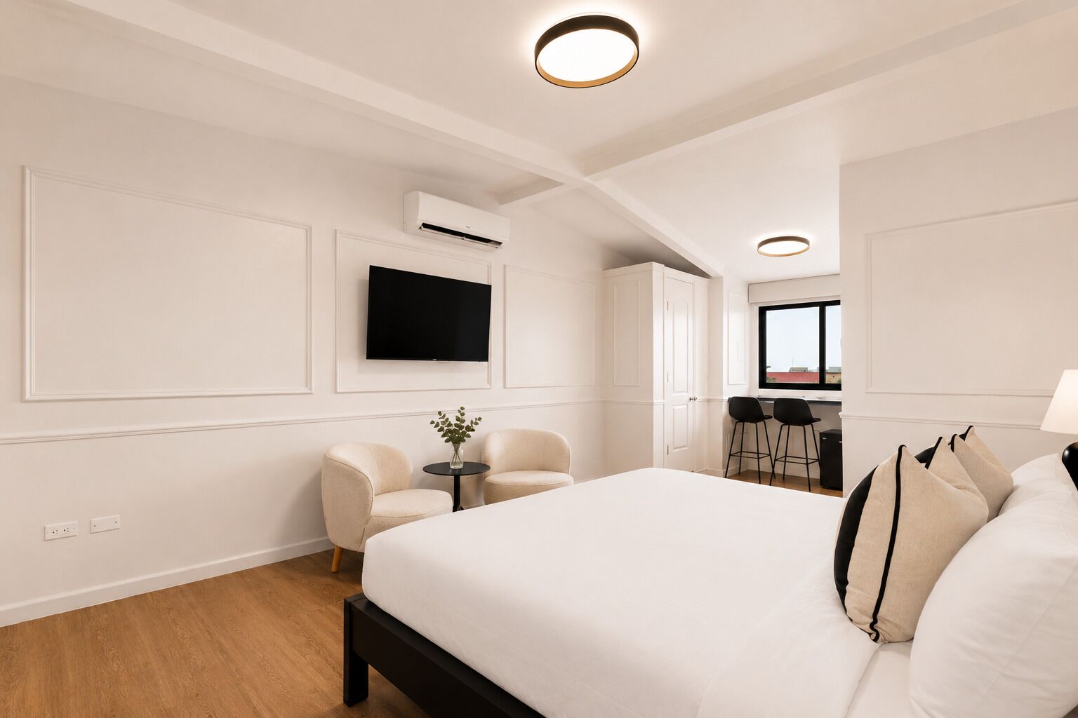 A modern hotel room with a large bed, a TV, a desk with two chairs, and an air conditioning unit.