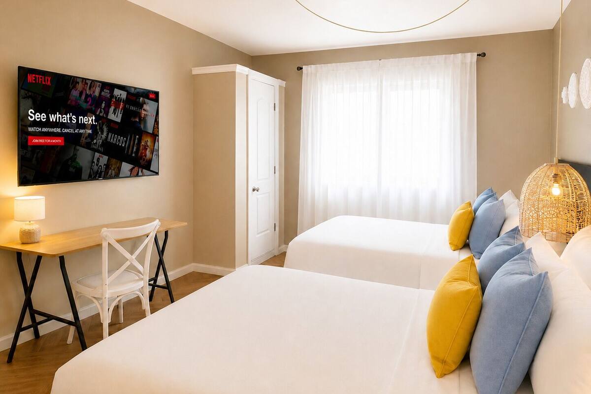 A hotel room with two beds, a desk with a chair, a television displaying Netflix content, and a window with curtains.