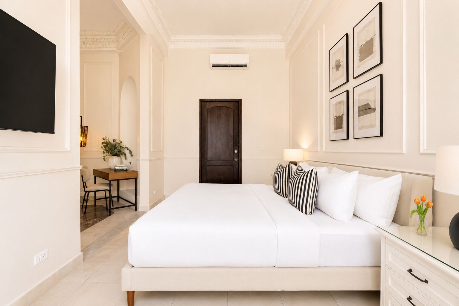 A hotel room with a bed, bedside table, lamp, framed pictures, and a desk with a chair.