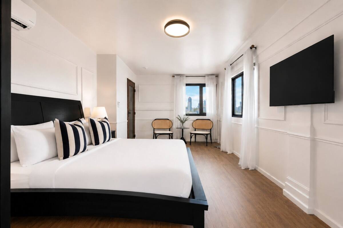 A hotel room with a large bed, a flat-screen TV mounted on the wall, a city view from the window, and a small table with chairs.