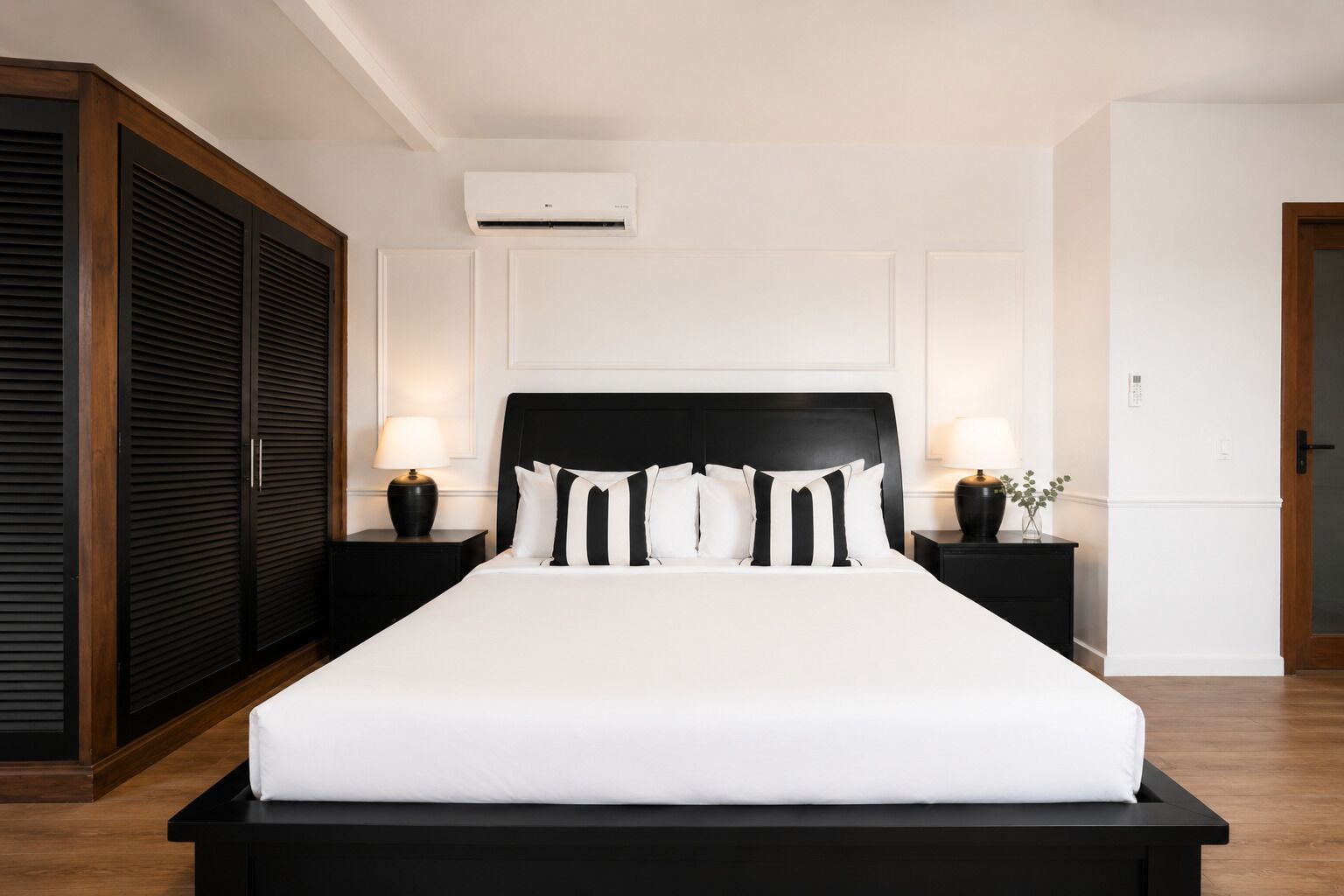 A hotel room with a large bed, two bedside tables with lamps, a wardrobe, and an air conditioning unit.