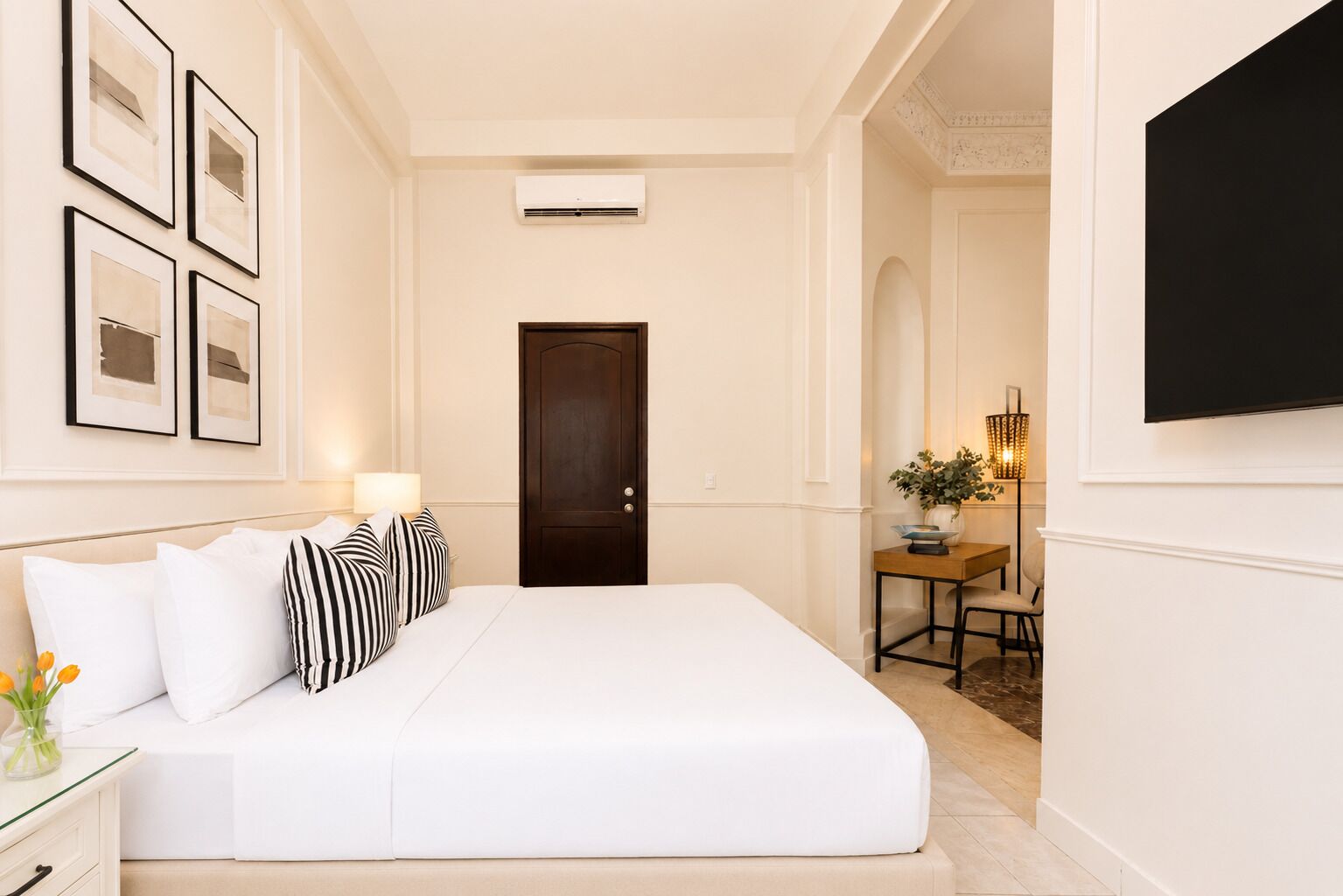 A hotel room with a bed, a desk with a lamp, a TV, and framed pictures on the wall.