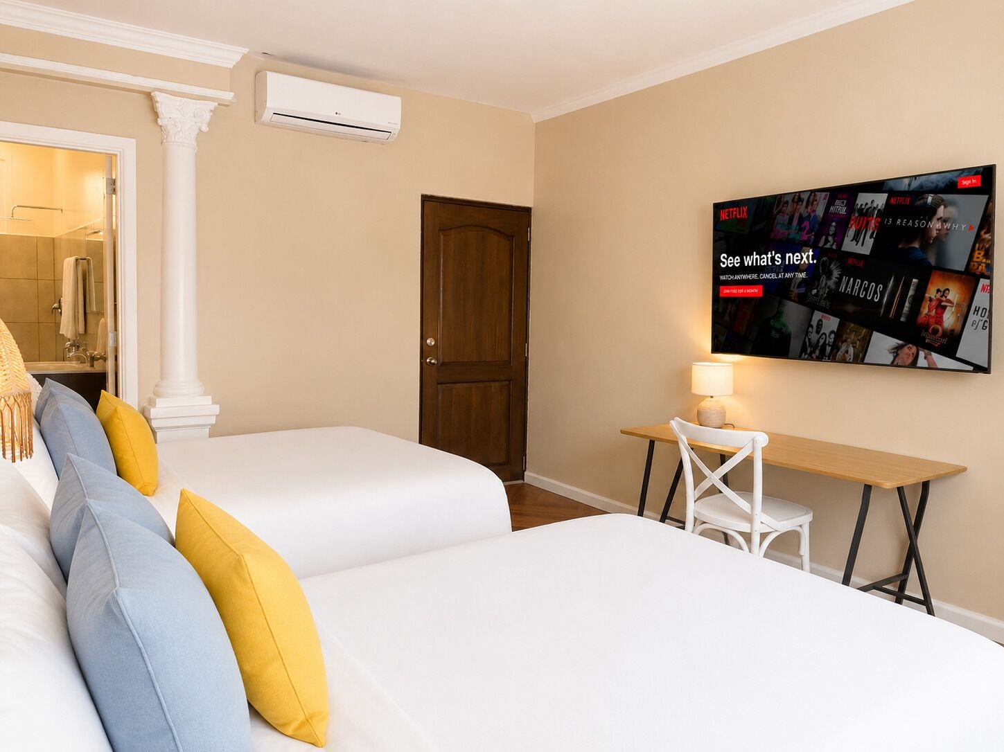 A hotel room with two beds, a desk, a chair, a TV displaying a Netflix interface, and an air conditioning unit.