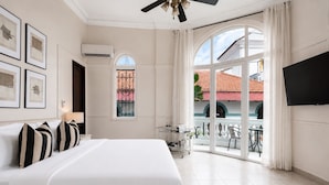 Ein Hotelzimmer mit einem großen Bett, einem Fernseher, einem Balkon mit Blick nach außen und einem Fenster mit schmiedeeisernem Zierwerk.