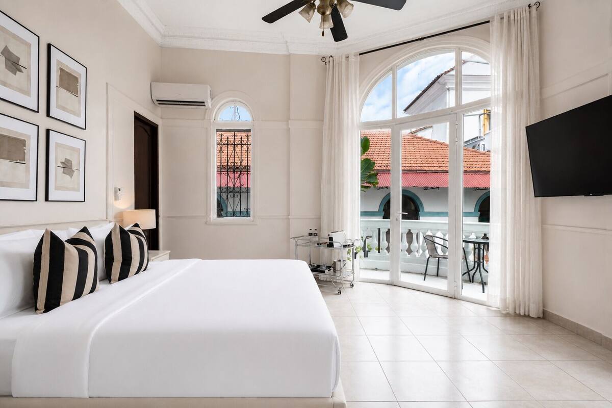 A hotel room with a large bed, a TV, a balcony with a view of the outside, and a window with decorative ironwork.