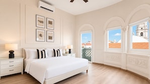 Ein Schlafzimmer mit einem großen Bett, weißen Wänden und Blick auf ein Gebäude durch Bogenfenster.