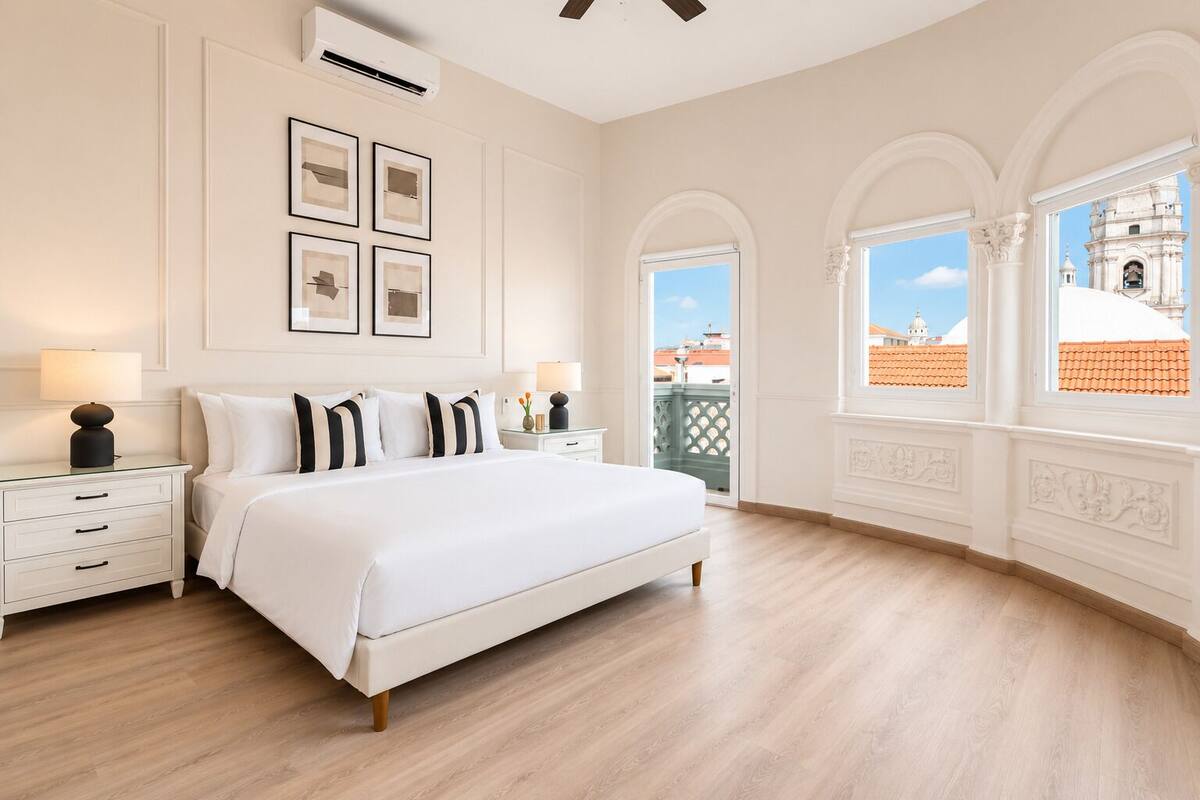 A bedroom with a large bed, white walls, and a view of a building through arched windows.